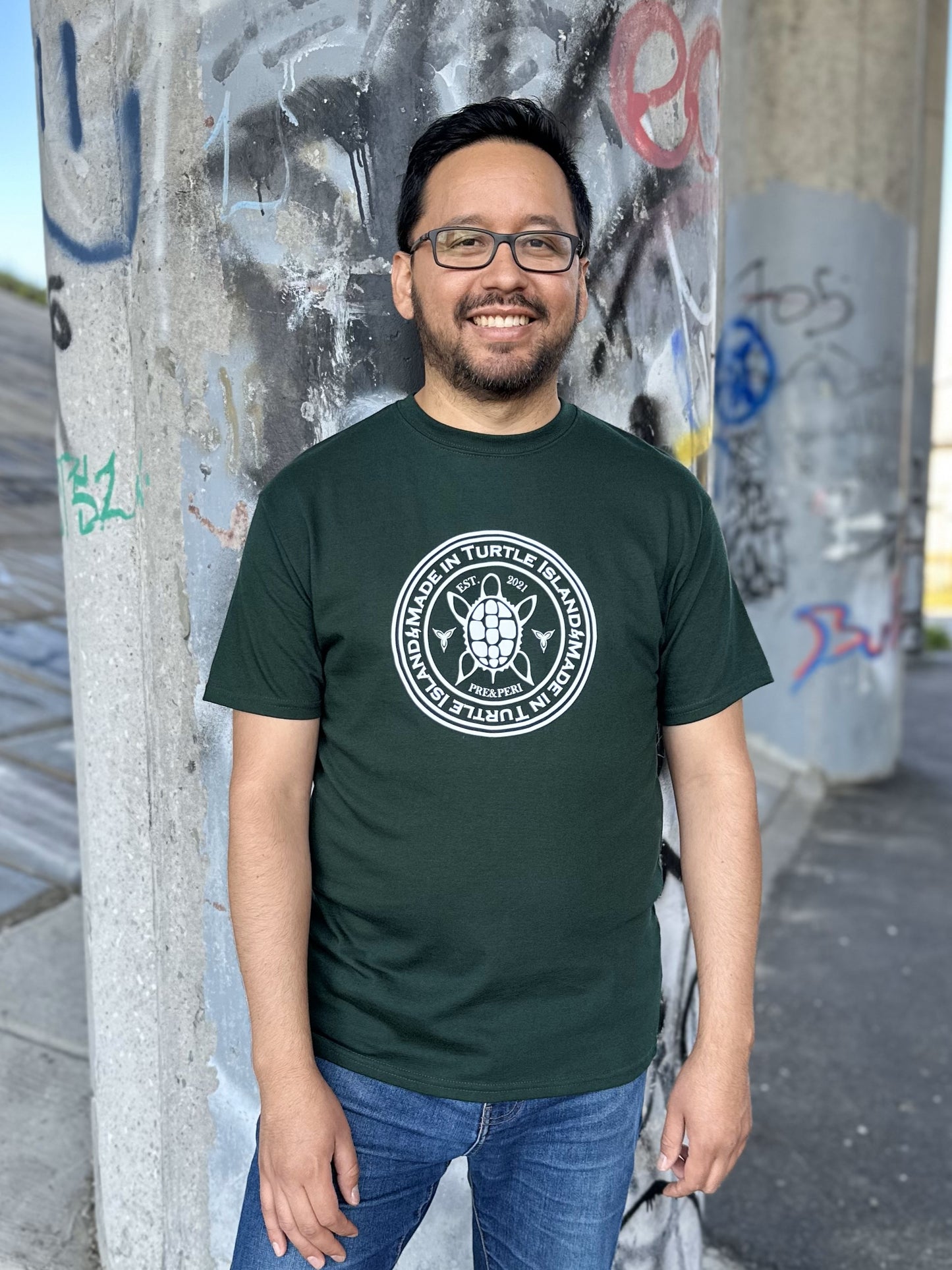A man wearing a forest green t-shirt with a white print of a turtle with the words "Made in Turtle Island" around it.