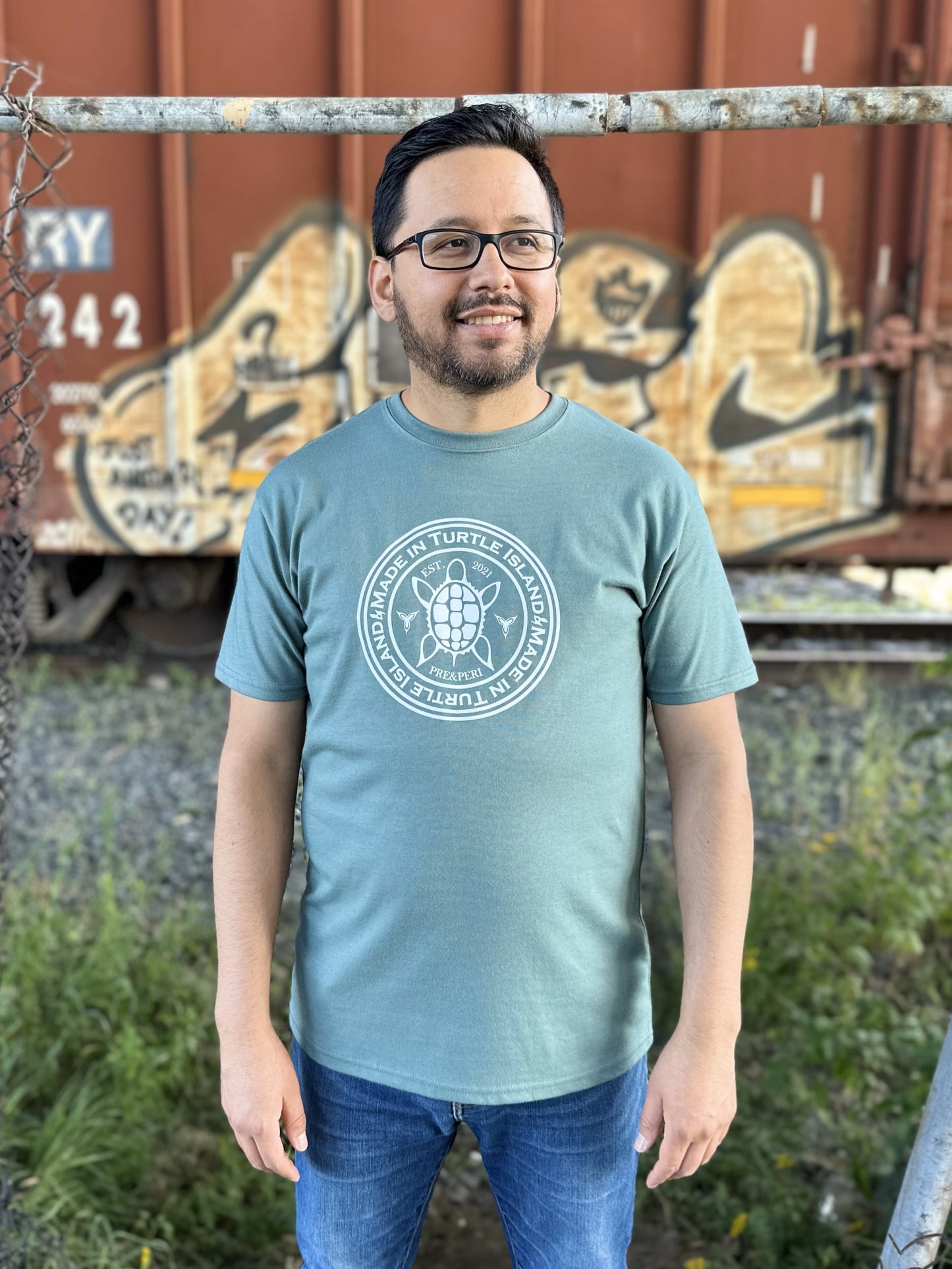 A man wearing a sage green t-shirt with a white print of a turtle with the words "Made in Turtle Island" around it.