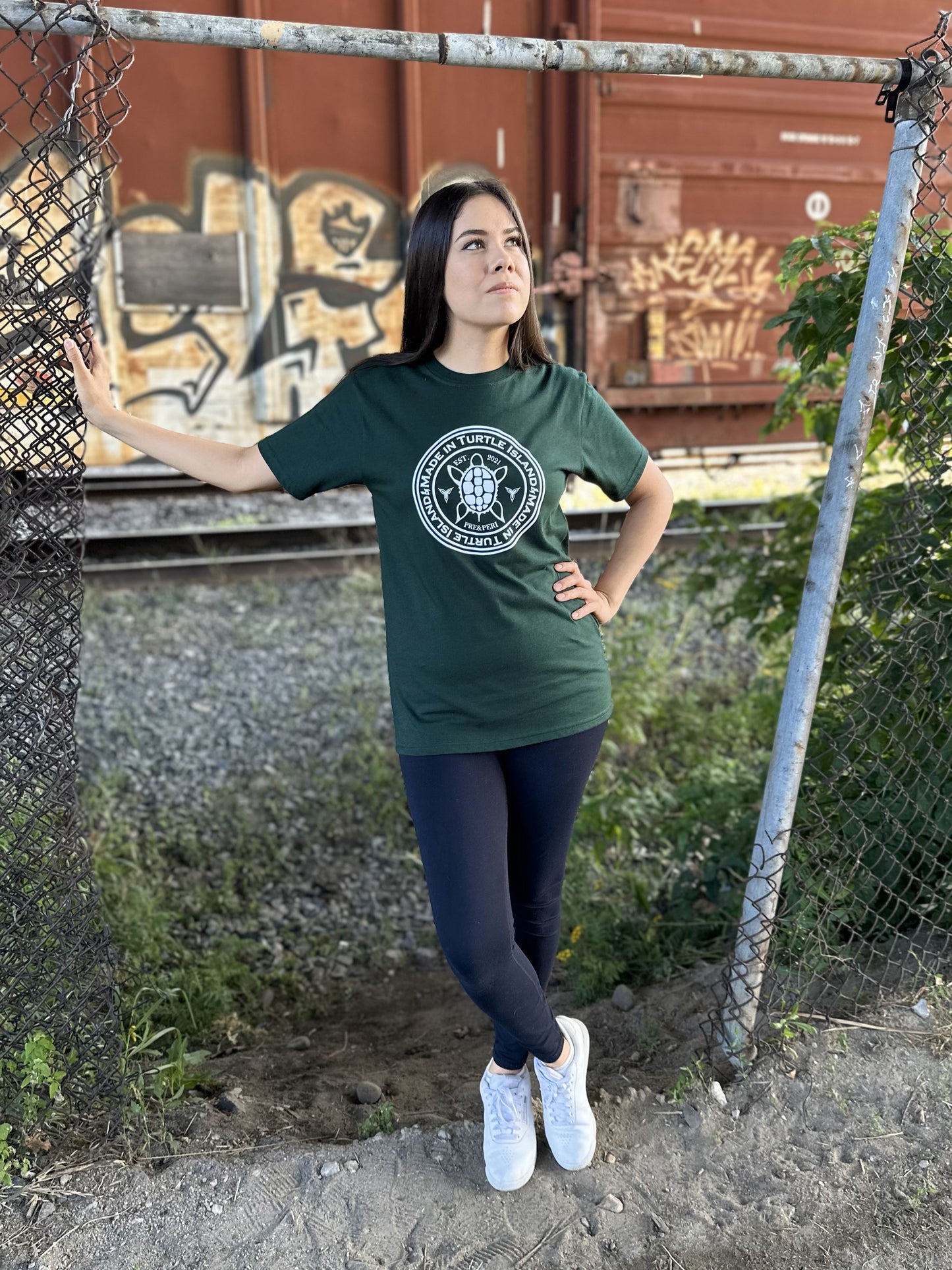 A woman wearing a forest green t-shirt with a white print of a turtle with the words "Made in Turtle Island" around it.
