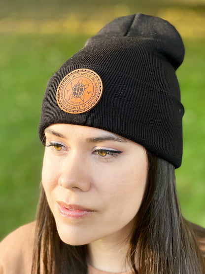 A woman wearing a black toque with a leather patch with and engraving of a turtle with the words "Made in Turtle Island" around it.
