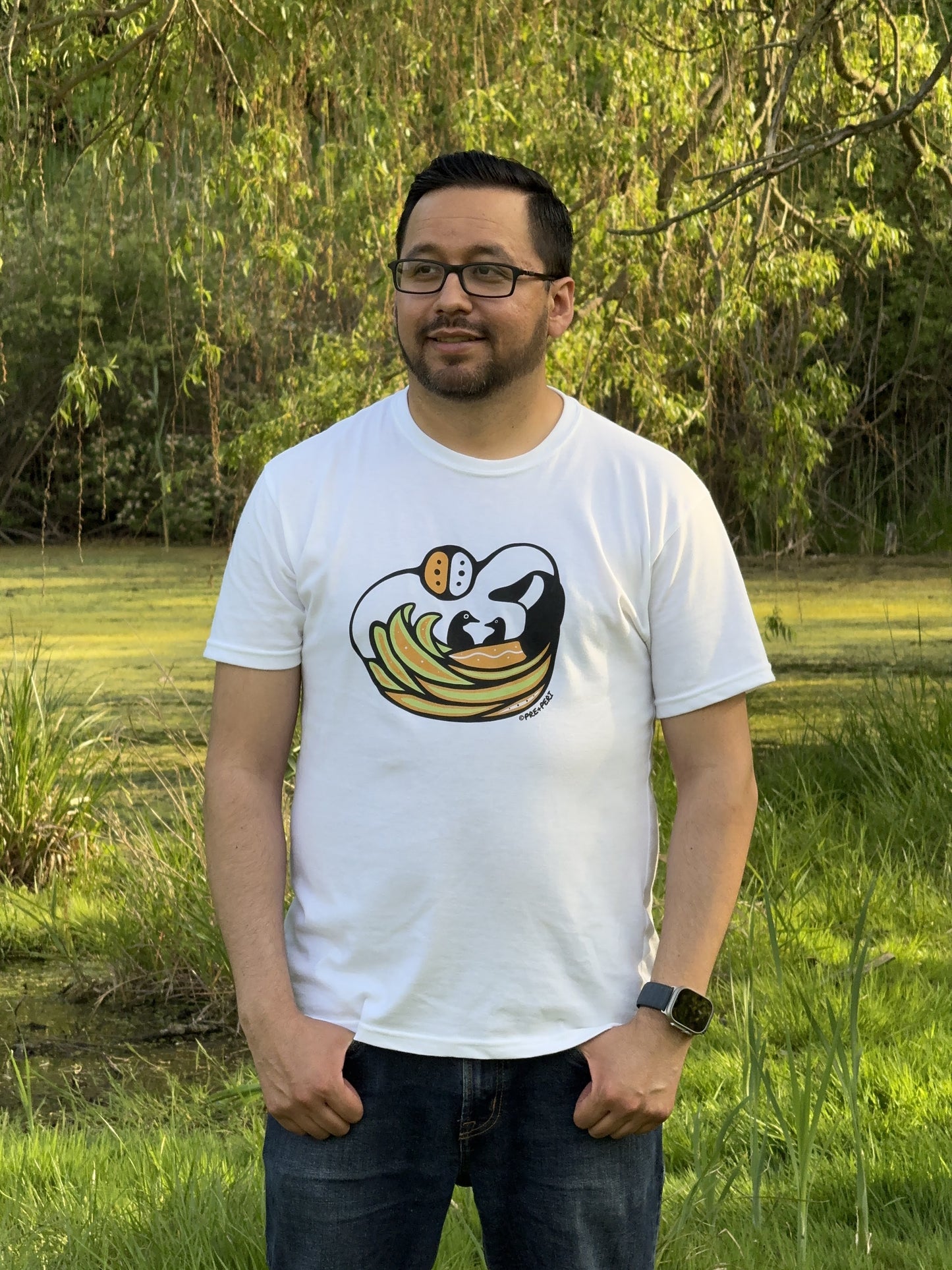 A man wearing a white t-shirt with a Woodland Art style Canada Goose design. The mother goose is keeping her two babies safe in her wings.