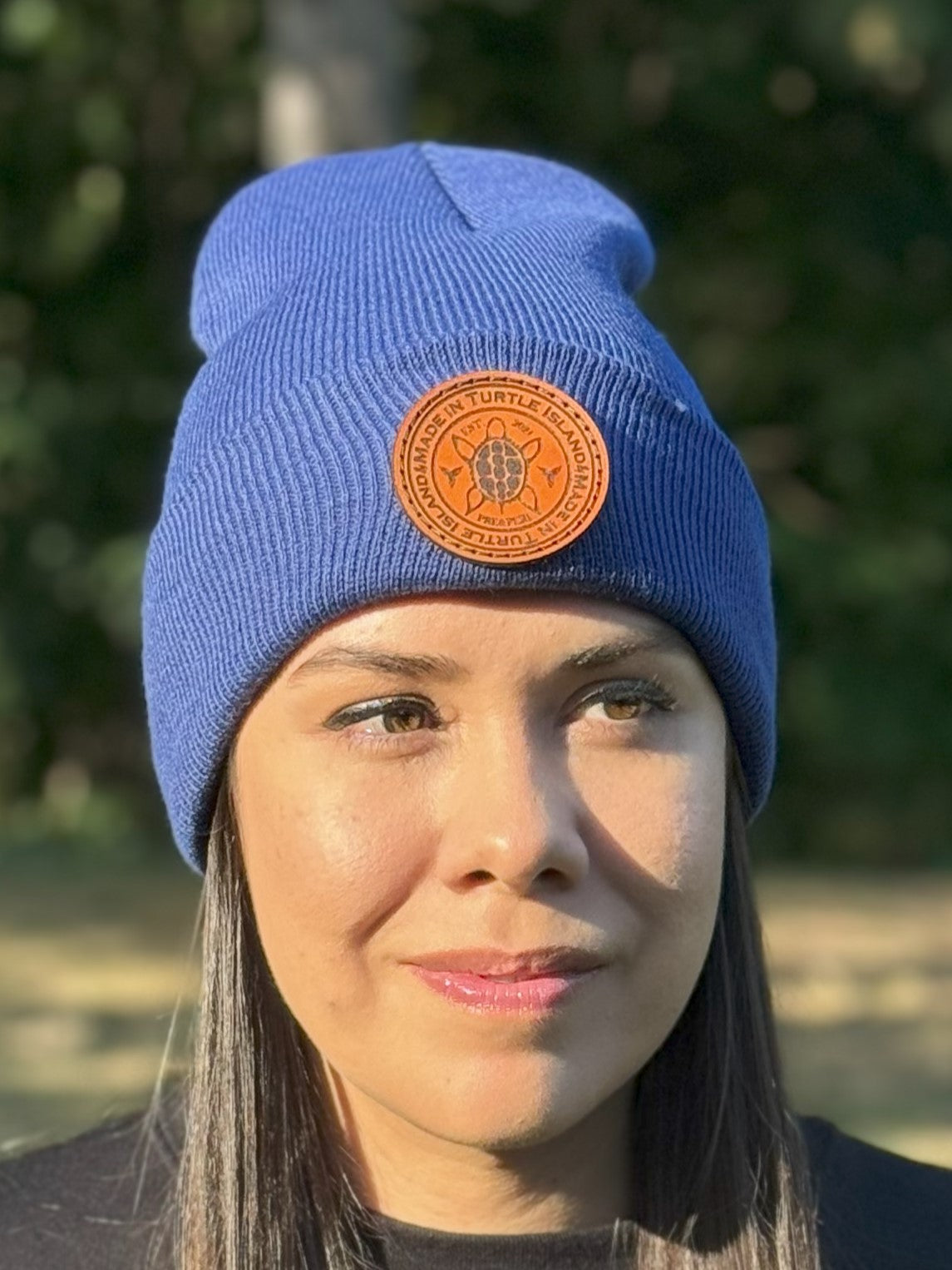 A woman wearing a blue acrylic rib knit toque with a leather patch engraved with a turtle with the words "Made in Turtle Island" around it.