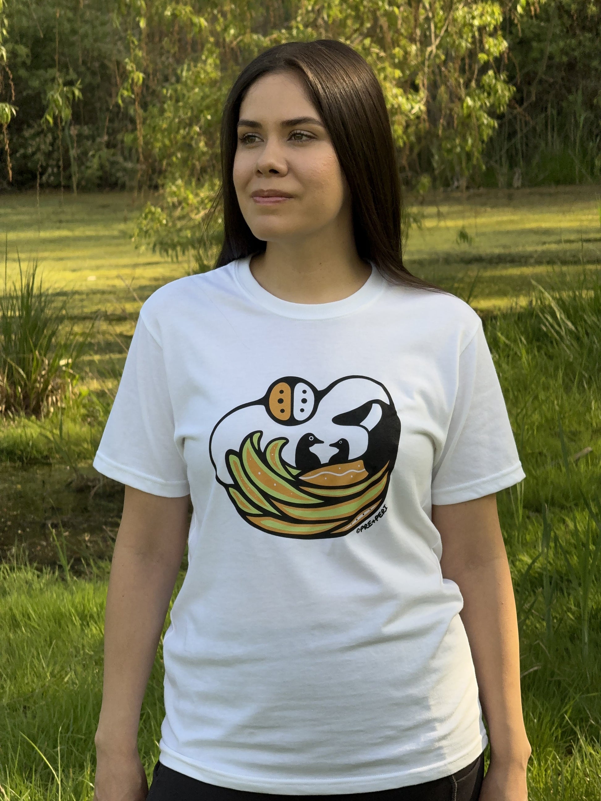 A woman wearing a white t-shirt with a Woodland Art style Canada Goose design. The mother goose is keeping her two babies safe in her wings.