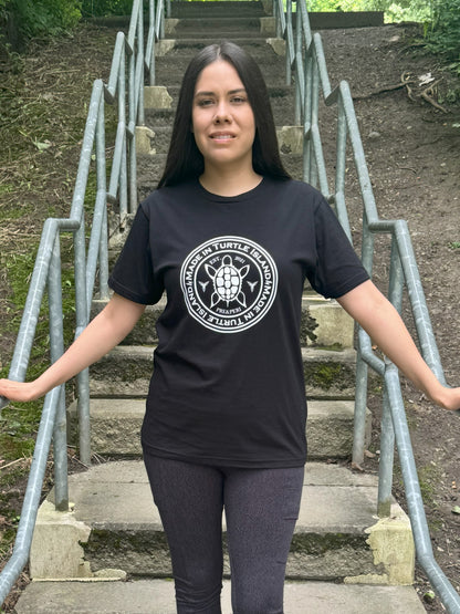 A woman standing on outdoor steps wearing a black organic cotton t-shirt with a white print that says "Made in Turtle Island" around a central image of a turtle.