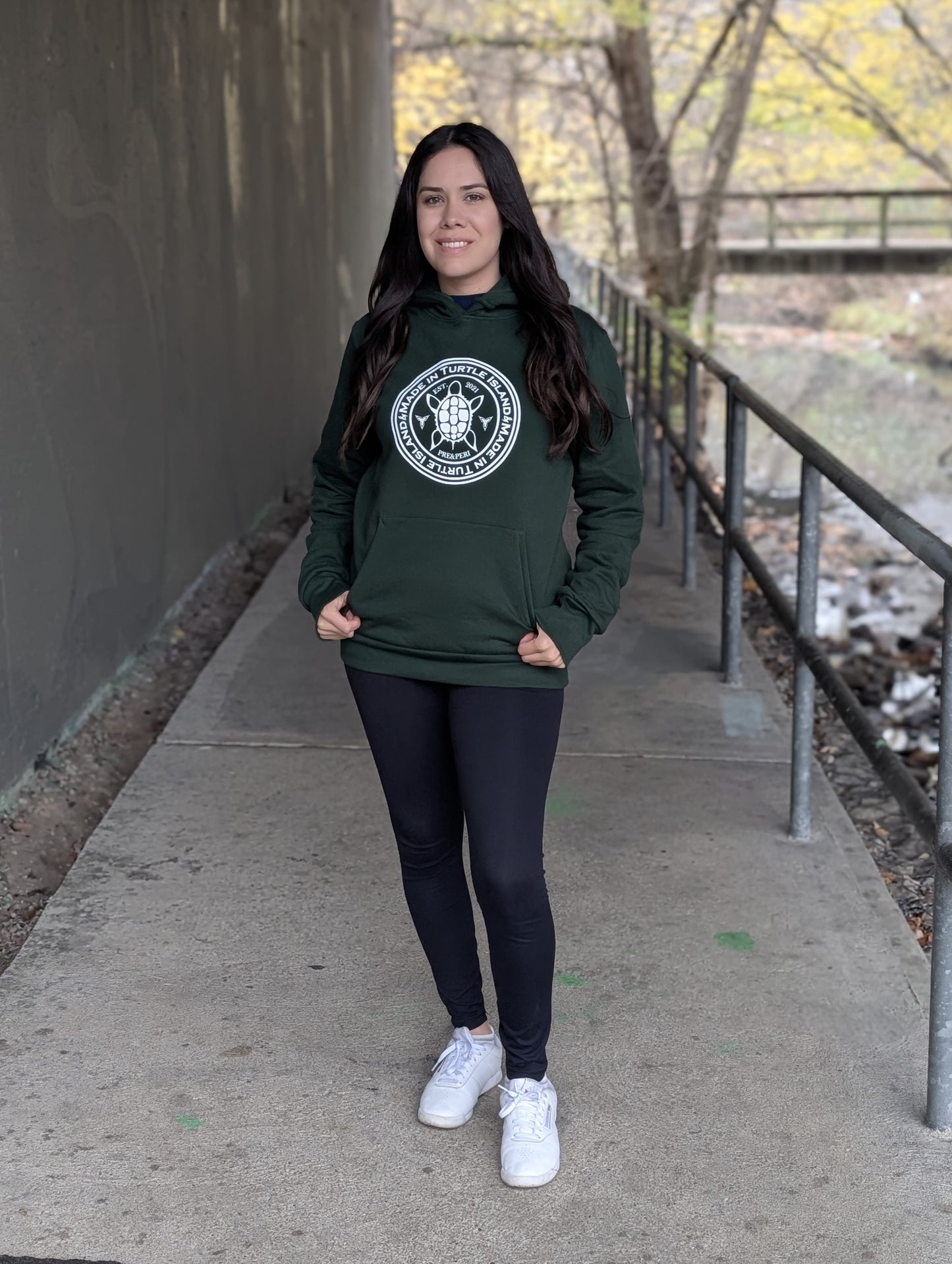 A woman wearing a forest green hoodie with kangaroo pocket. White artwork of a stylized turtle with the words "Made in Turtle Island" around it.