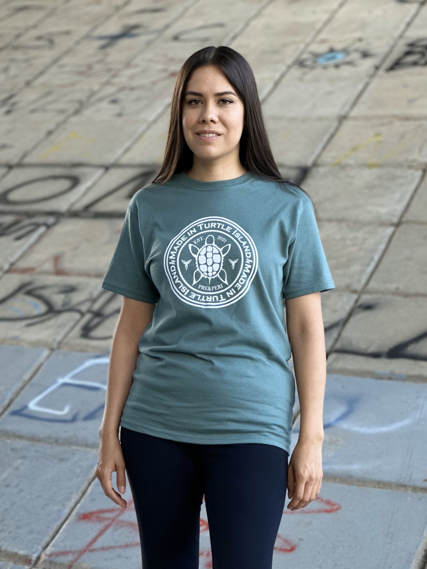 A woman wearing a sage green t-shirt with a white print of a turtle with the words "Made in Turtle Island" around it.