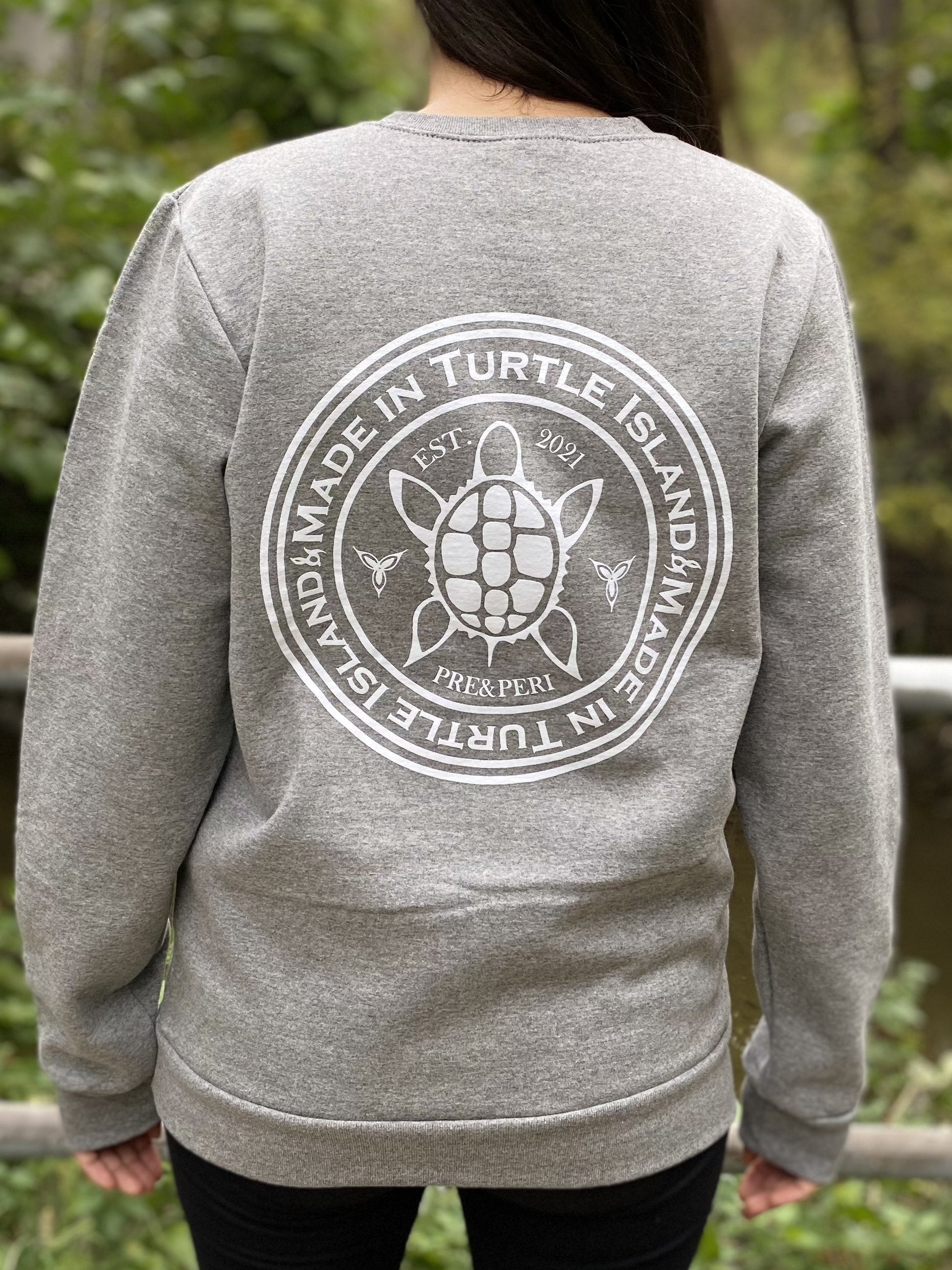 Back of a grey heather sweatshirt on a woman with a white print of a turtle in the centre of a circle with words "Made in Turtle Island" around it twice