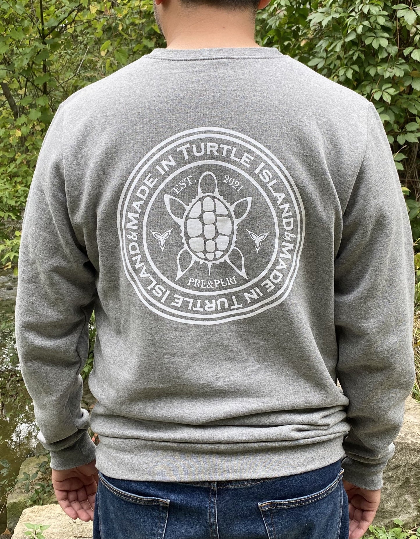 Back of a grey heather sweatshirt on a man with a white print of a turtle in the centre of a circle with words "Made in Turtle Island" around it twice