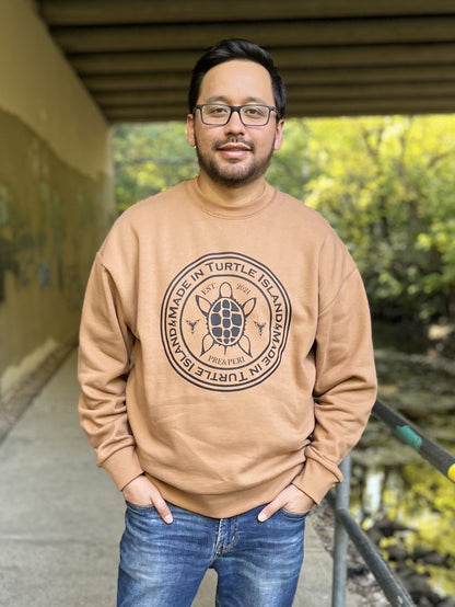 A man wearing a camel-colour oversized crewneck sweatshirt with a black print depicting a stylized turtles flanked by two trilliums all in a circle with the words "Made in Turtle Island" in a circle