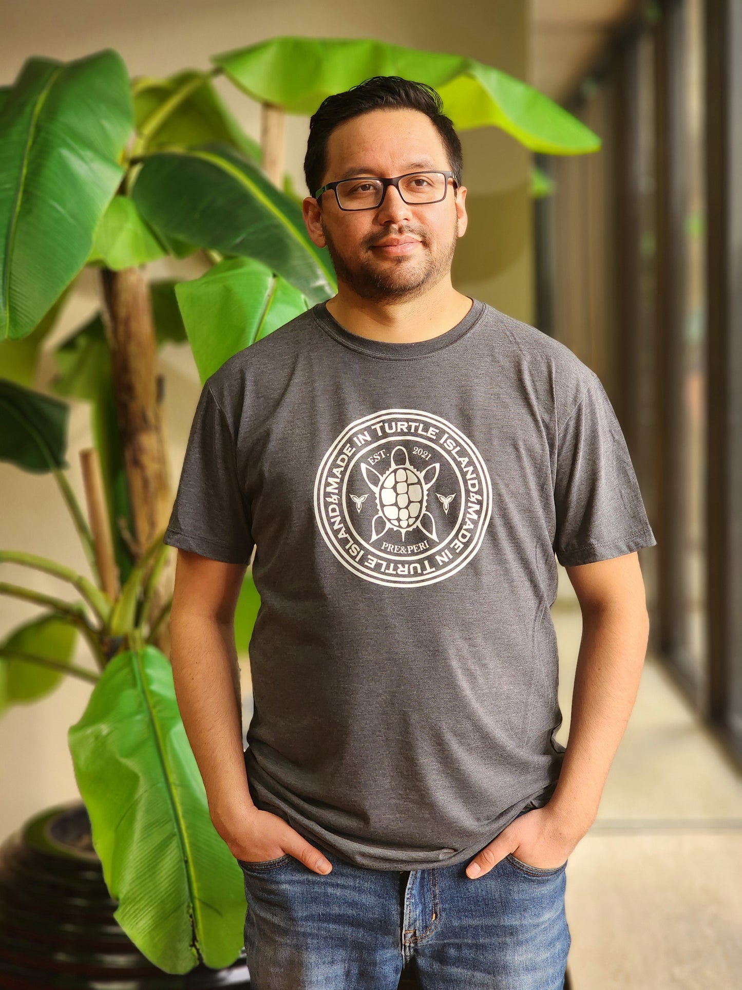 A man wearing a heather charcoal t-shirt with a white print of a turtle in the centre of a circle with words "Made in Turtle Island" around it twice