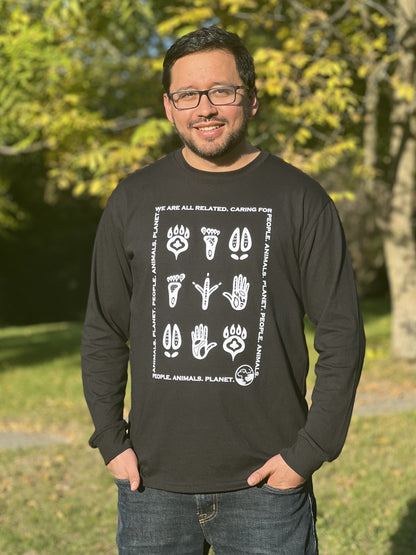 A man wearing a long-sleeved black t-shirt with a white print that is made up of Indigenous-style footprints of animals and humans with the words "We are all related. Caring for People. Animals. Planet." in a rectangular shape around the prints.