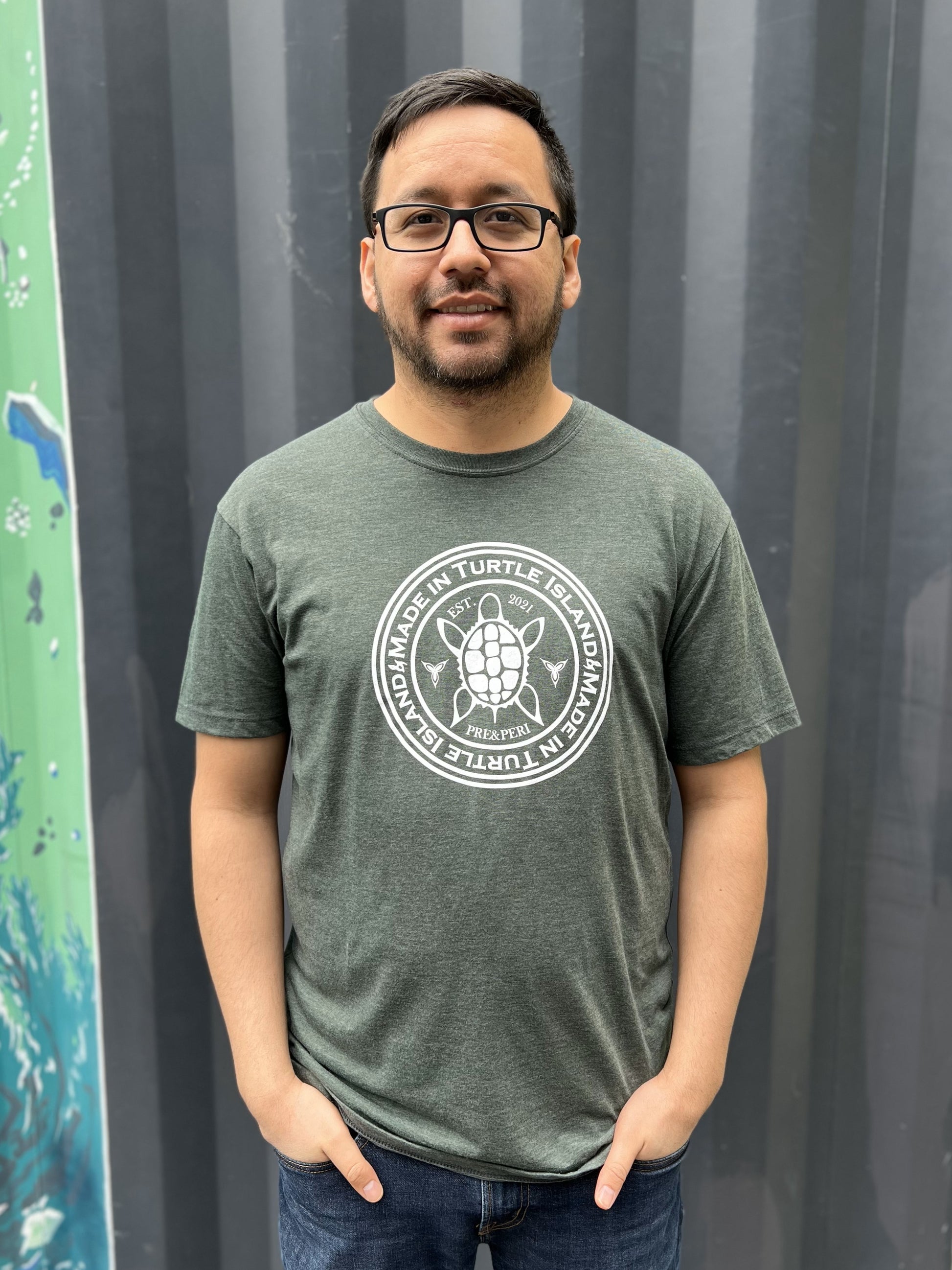 A man wearing a forest green heather t-shirt with a white print of a turtle in the centre of a circle with words "Made in Turtle Island" around it twice