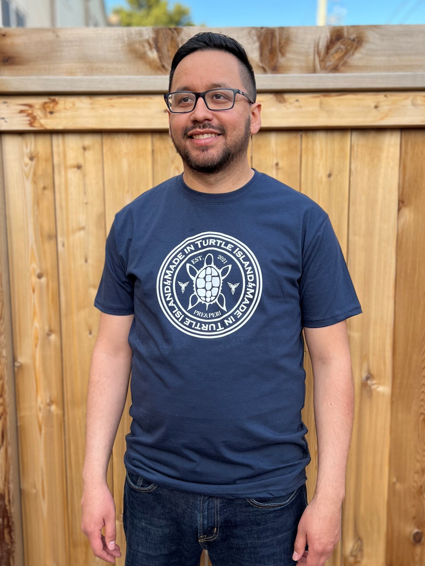 A man wearing a navy unisex t-shirt with a white print of a Turtle flanked by trilliums and with the words "Made in Turtle Island" around it in a circle. 
