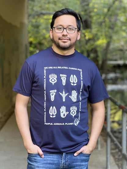 A man wearing a midnight blue t-shirt with a white print of stylized animal and human footprints encased by a rectangle formed by the words "Wea are all related. Caring for People. Animals, Planet."