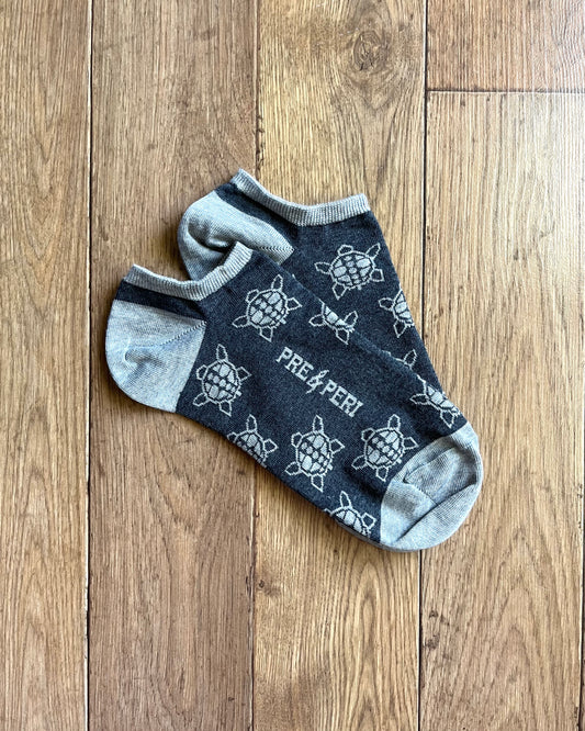 A photo of a pair of ankle socks on a wood flooring background. The socks are dark grey with light grey heel, toe, cuff and repeated turtle design throughout.