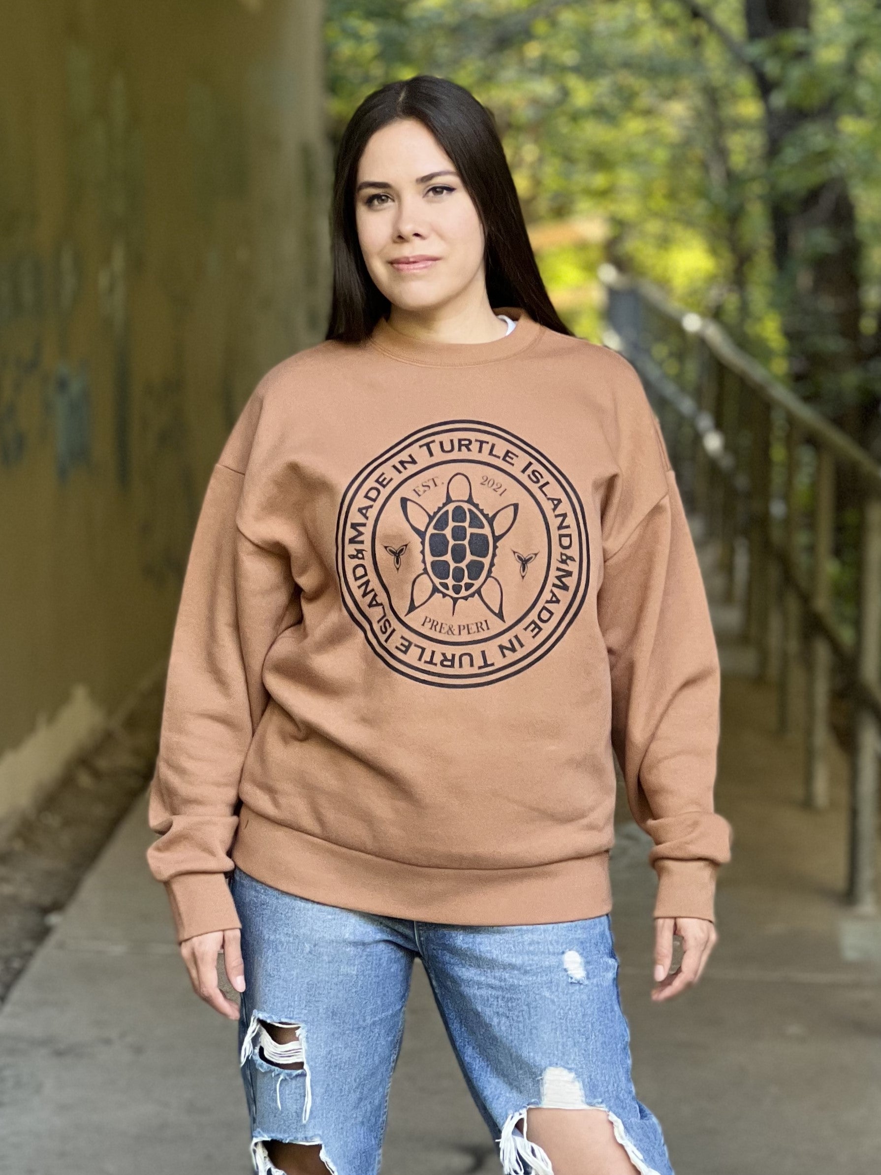 A woman wearing a camel-colour oversized crewneck sweatshirt with a black print depicting a stylized turtles flanked by two trilliums all in a circle with the words "Made in Turtle Island" in a circle