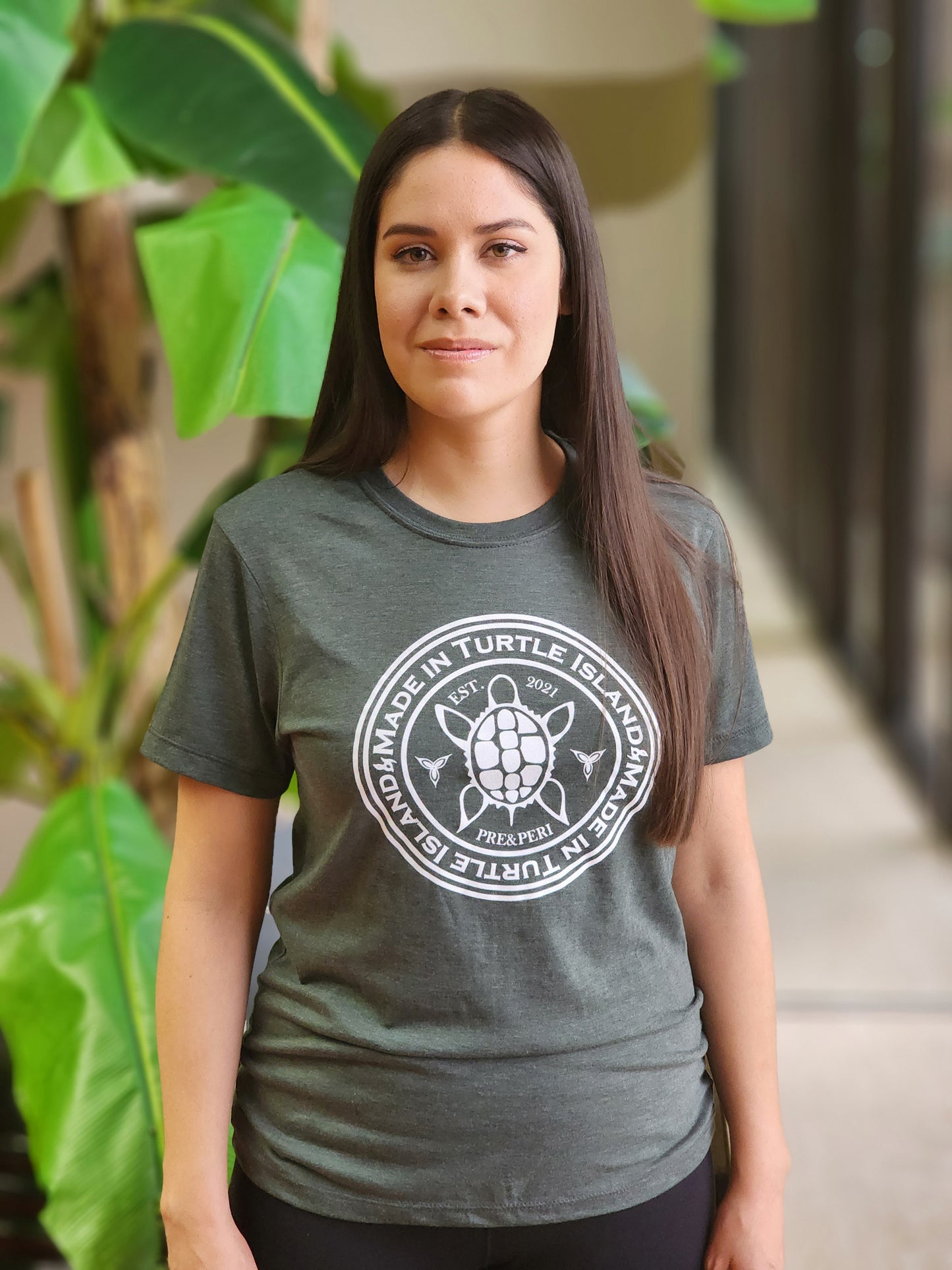 A woman wearing a forest green heather t-shirt with a white print of a turtle in the centre of a circle with words "Made in Turtle Island" around it twice