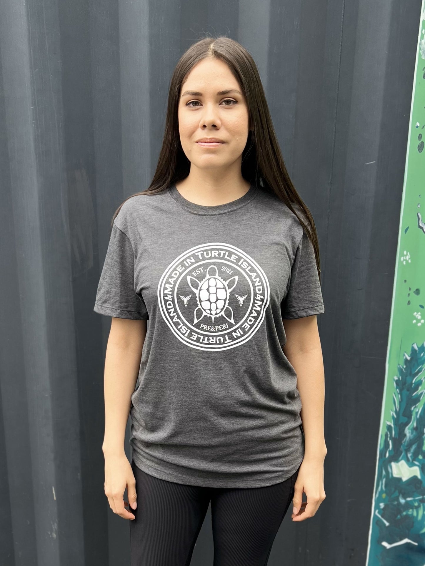 A woman wearing a heather charcoal t-shirt with a white print of a turtle in the centre of a circle with words "Made in Turtle Island" around it twice