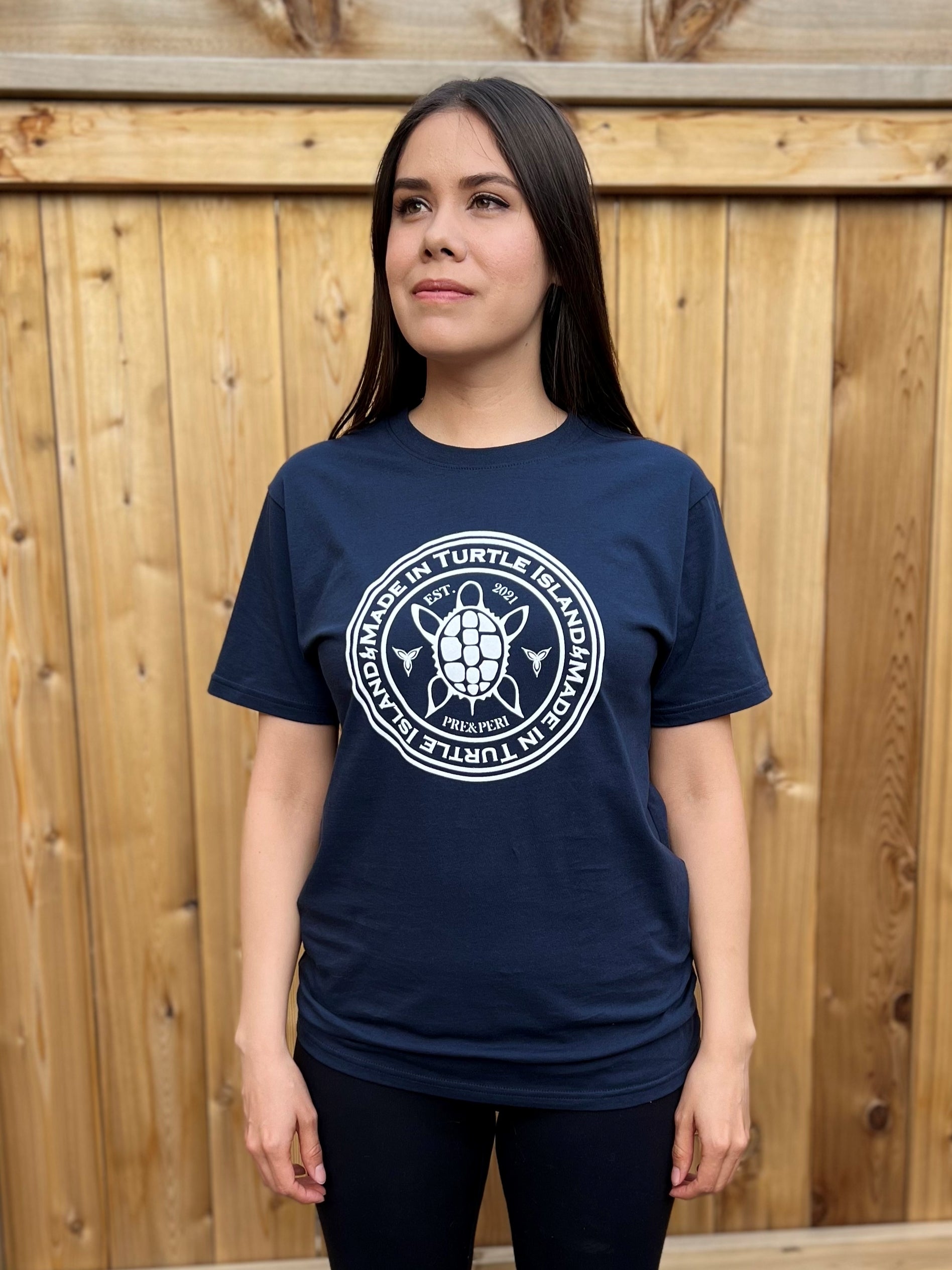A woman wearing a navy unisex t-shirt with a white print of a Turtle flanked by trilliums and with the words "Made in Turtle Island" around it in a circle. 