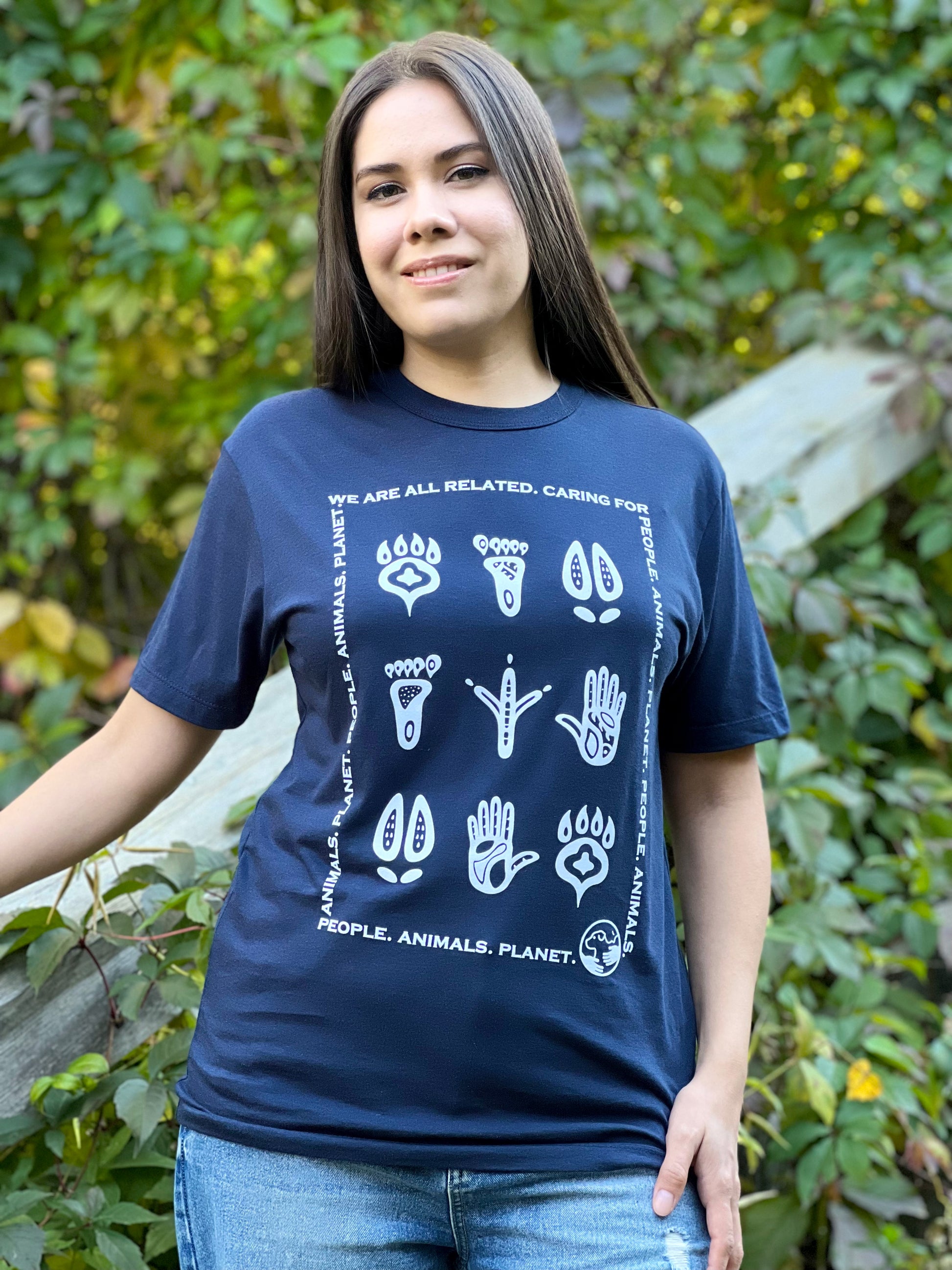 A woman wearing a midnight blue t-shirt with a white print of stylized animal and human footprints encased by a rectangle formed by the words "Wea are all related. Caring for People. Animals, Planet."
