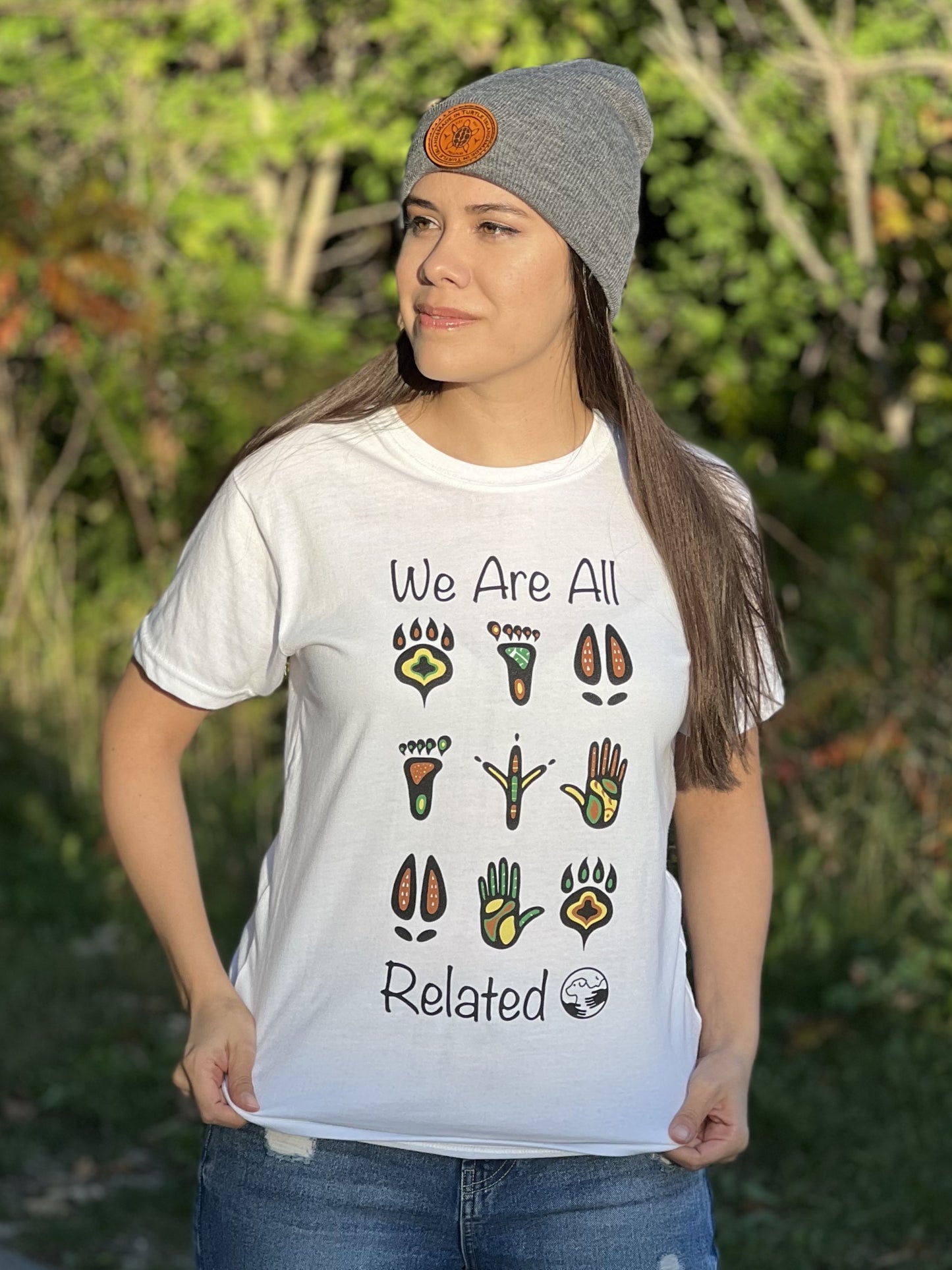 A woman wearing a white t-shirt with a mutlti-coloured print of stylized animal and human footprints. The words "We are all" are above the footprints "Related" is below the footprints with the One Health Partners logo beside. 