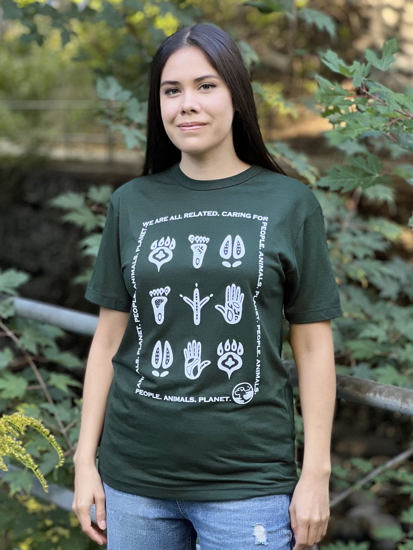 A woman wearing a forest green t-shirt with a white print of stylized animal and human footprints encased by a rectangle formed by the words "We are all related. Caring for People. Animals, Planet."