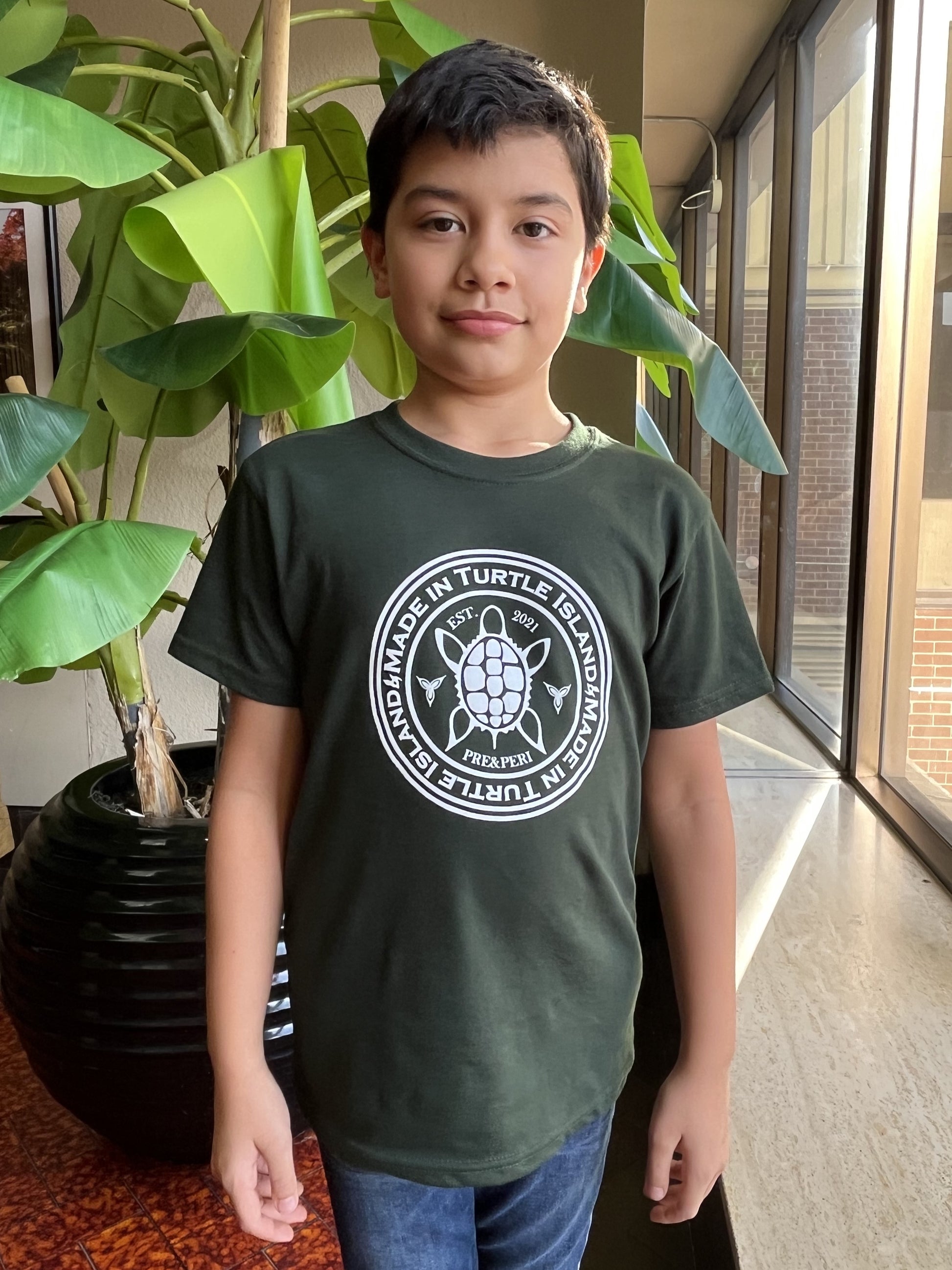 A boy wearing a forest green t-shirt with a white print of a turtle in a circle with the words "Made in Turtle Island" around it.