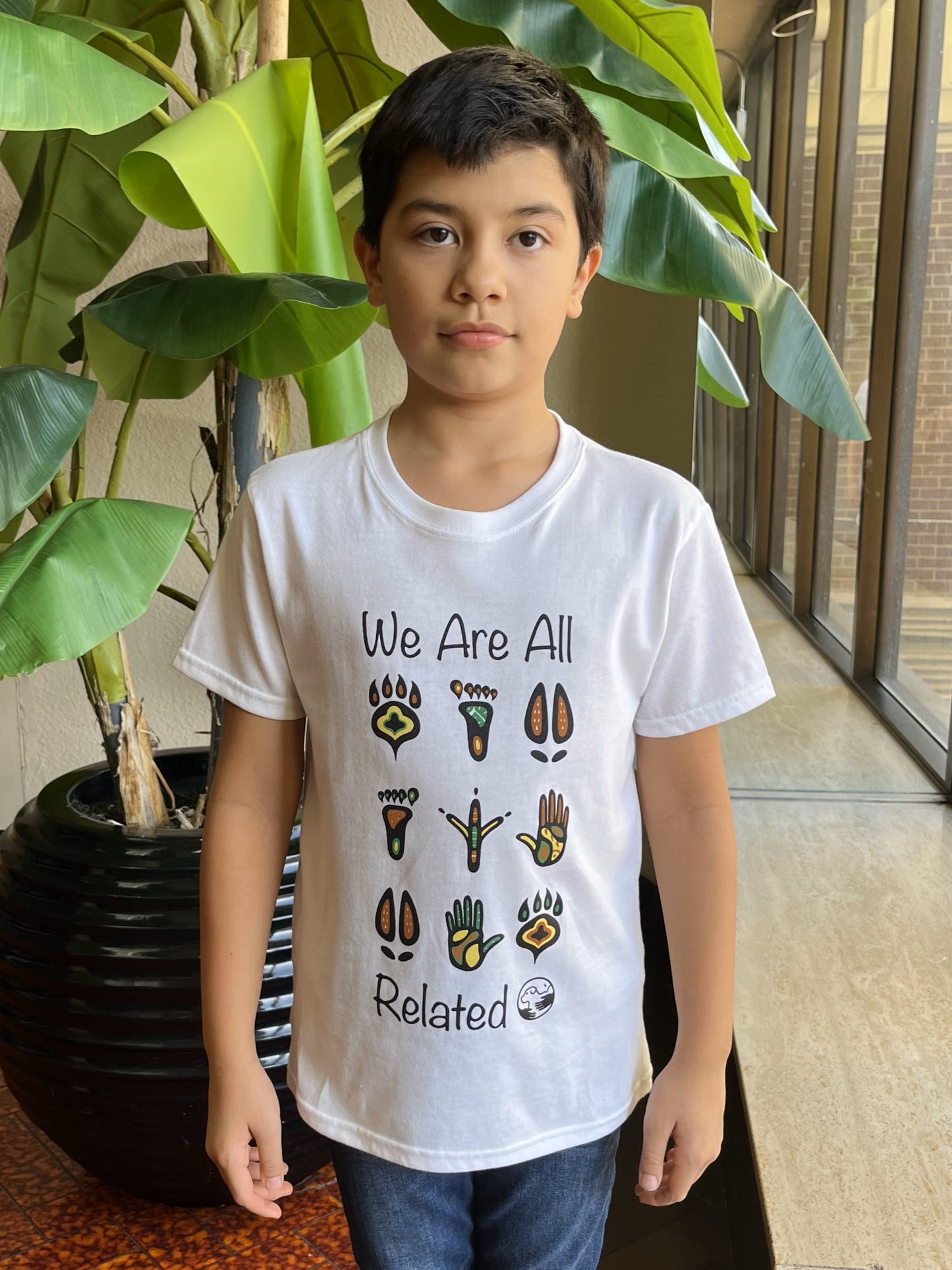 A boy wearing a white t-shirt with the words "We Are All Related" and multi-colour Indigenous-style animal and human footprints.