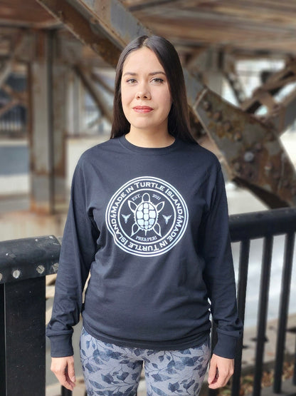 A woman wearing a black long sleeve t-shirt with a white print of a turtle in the centre of a circle with words "Made in Turtle Island" around it twice