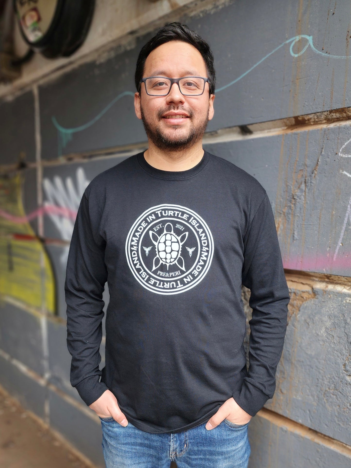A man wearing a black long sleeve t-shirt with a white print of a turtle in the centre of a circle with words "Made in Turtle Island" around it twice